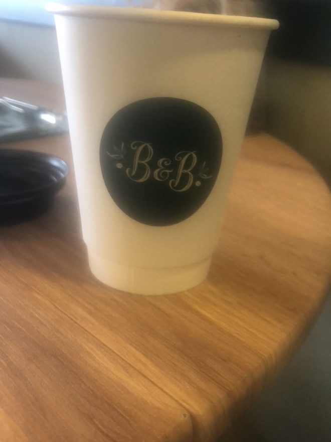 A takeaway coffee cup standing on a bare wooden table in warm light.